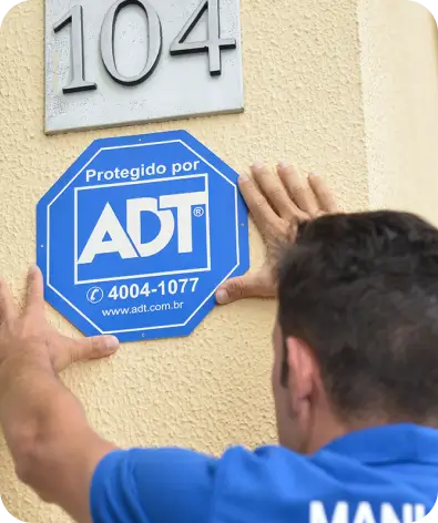 Imagem de um técnico da ADT colando uma placa da ADT na parade